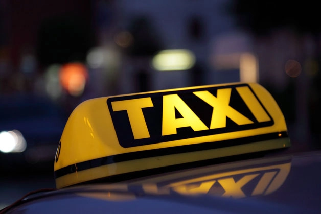 A bright yellow taxi sign glows atop a car in the evening. The bold black "TAXI" lettering is illuminated, set against a blurred city background.
