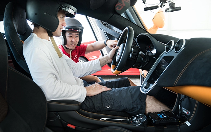 Two people in helmets sitting inside a race car. The driver, in a white shirt, listens attentively to the passenger explaining controls. The scene conveys focus and instruction.