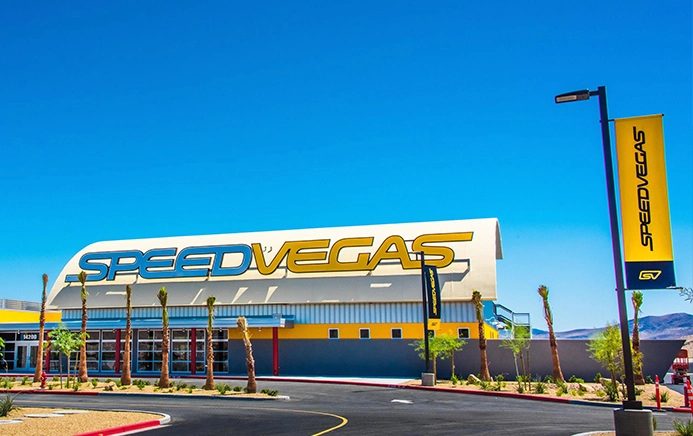 Modern building with "Speed Vegas" signage against a clear blue sky. Tall palm trees line the entrance. Bright yellow and blue theme creates an energetic vibe.