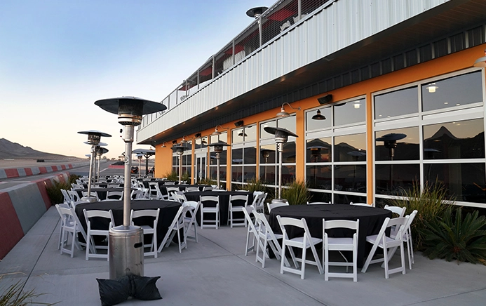 Outdoor patio with round tables set with black tablecloths and white chairs. Surrounded by tall patio heaters against a building with large windows. Desert landscape in the background.