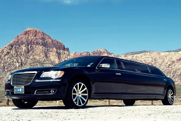 A sleek black limousine parked on a paved area against a backdrop of rugged, sunlit mountains under a clear blue sky, conveying luxury and grandeur.